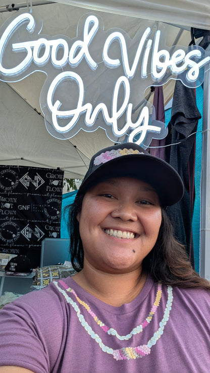 Person wearing a black foam trucker hat with Kahelelani shell design and a purple shirt with Ni'ihau shell lei design. 'Good Vibes Only' neon sign in the background. 