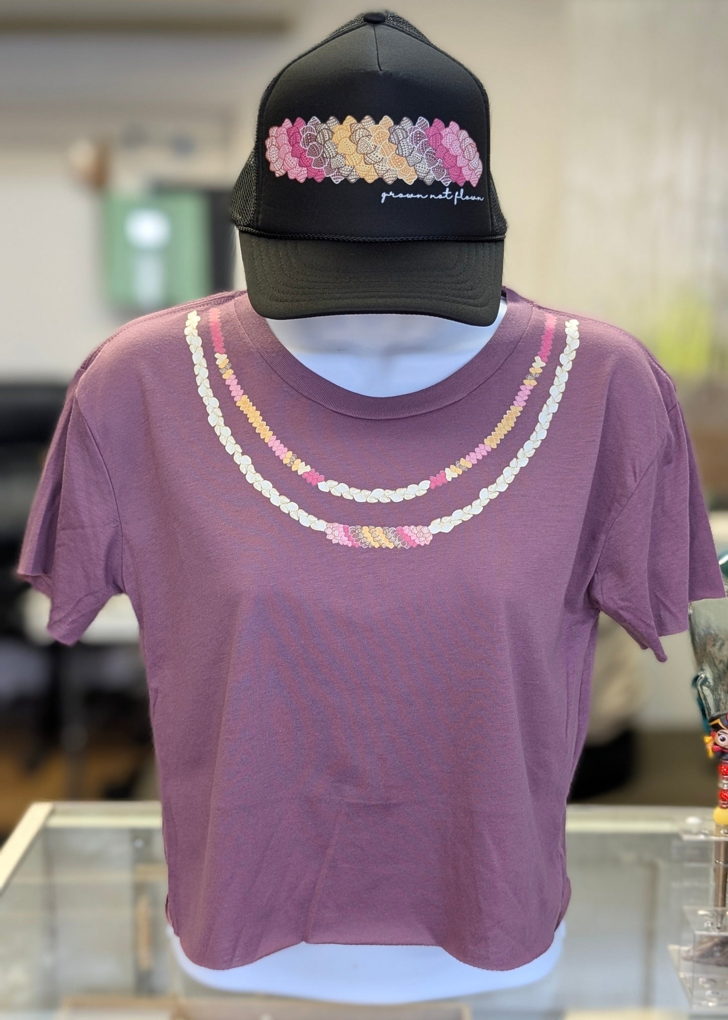 Mannequin wearing a purple cropped t-shirt with multi-color Ni'ihau shell lei design and a black foam trucker hat with multi-color kahelelani shell design.