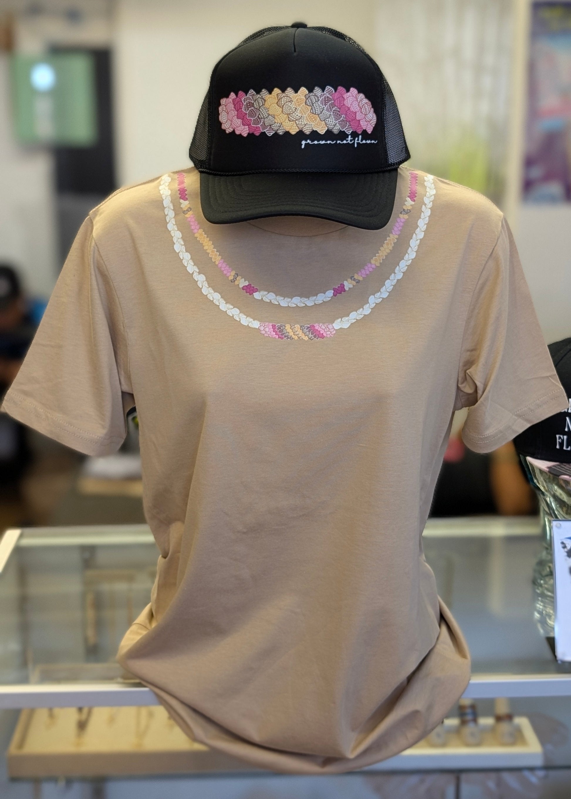 Beige t-shirt with decorative shell lei design and a black cap with colorful shell design on a mannequin.